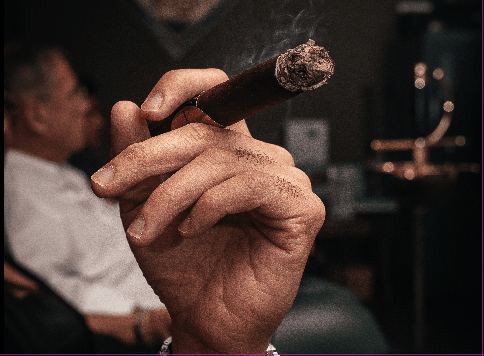 Close-up of a hand holding a lit cigar, with smoke rising.