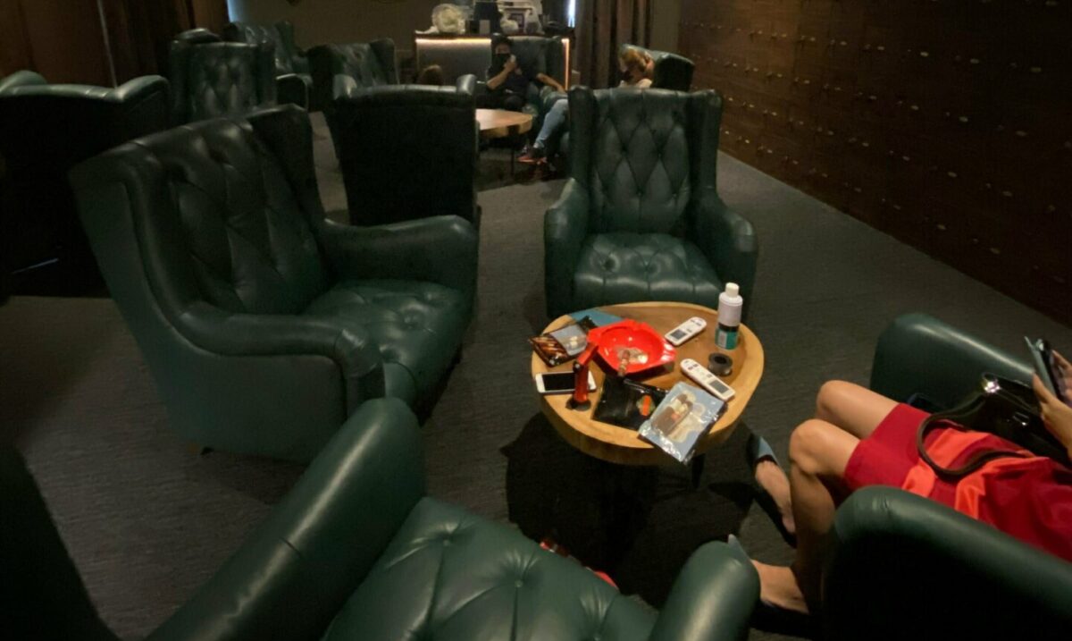 Dark cigar lounge with green leather wingbacks, humidor lockers on the wall, and a table cluttered with cutters, ashtray, remotes, and gear. Guests relax in the back and foreground.