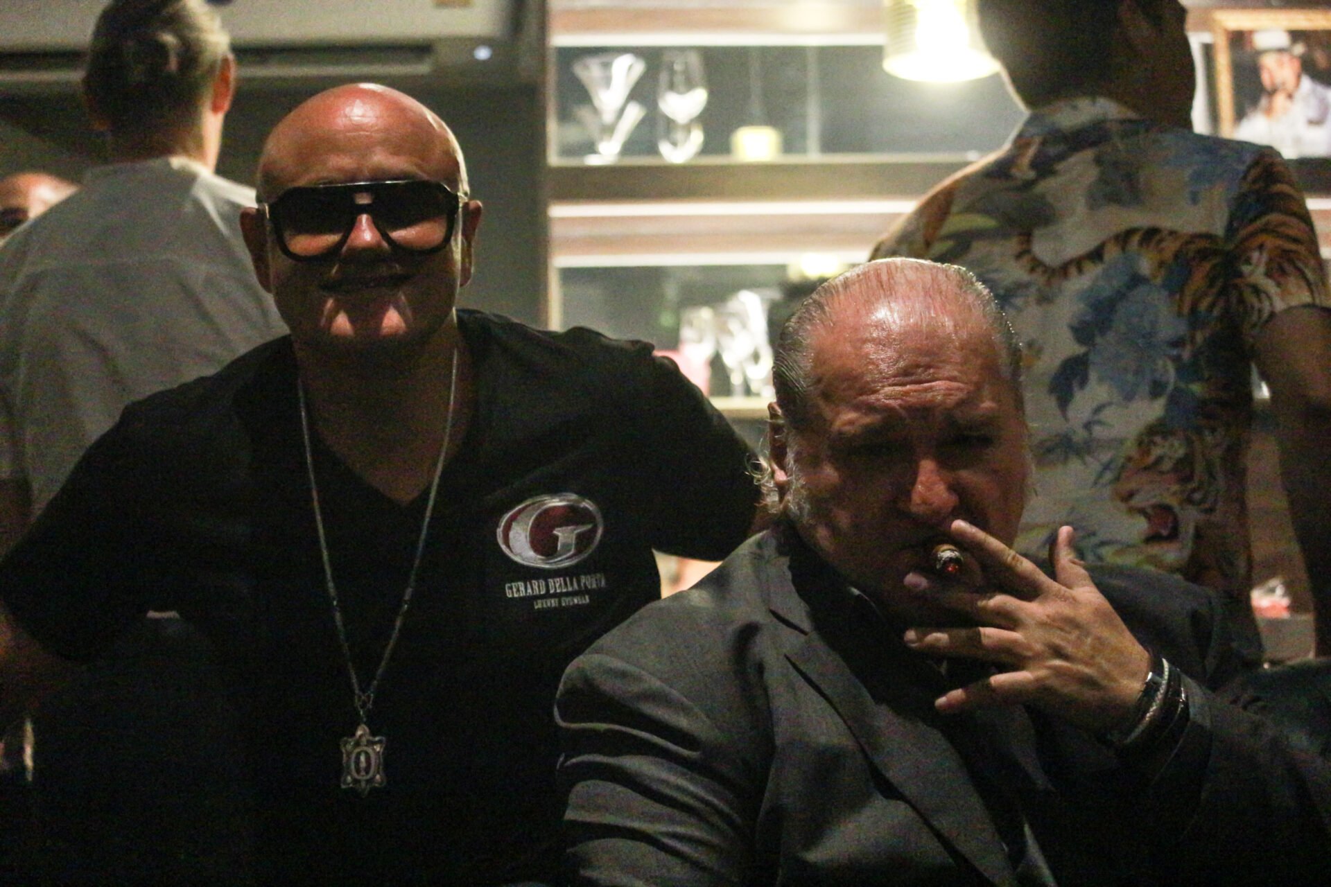 A bald man in sunglasses and a black t-shirt smiles at the camera, next to another man in a dark suit smoking a cigar, in a dimly lit lounge. Dr. Matthew Nekvapil is partially visible in the background.