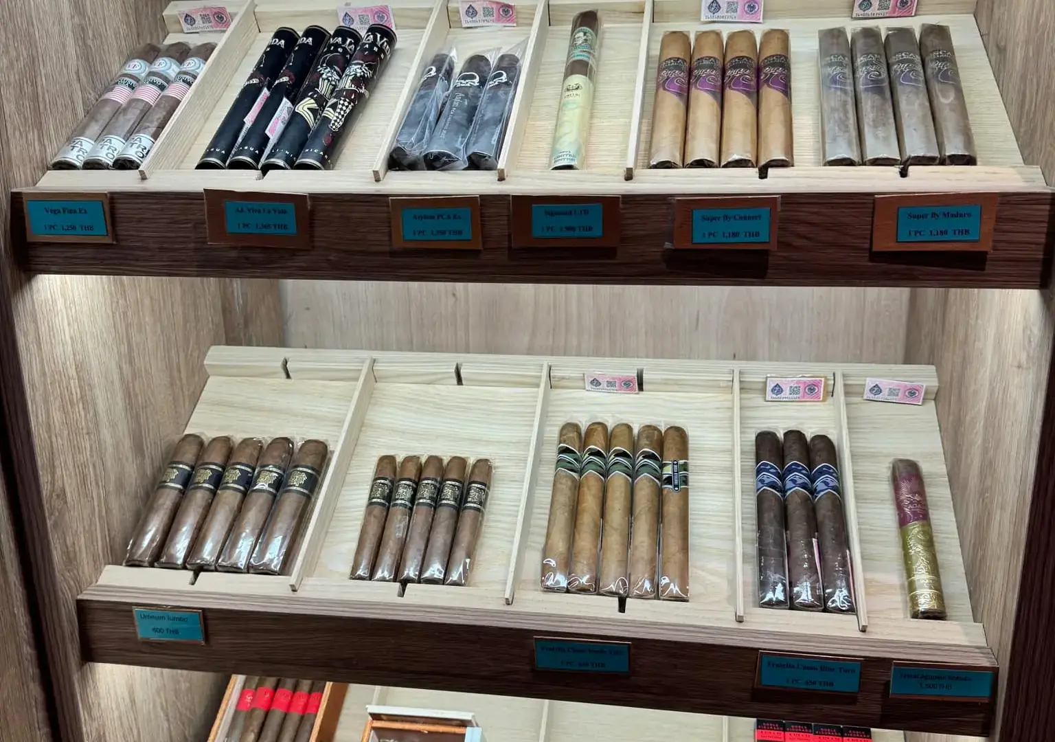 Close-up view of a wooden cigar display case shelf, showing multiple rows of individually priced premium cigars with various wrapper shades and sizes.