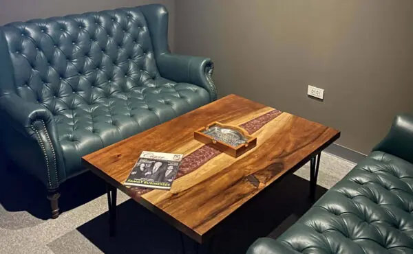 Interior of a private cigar room at MOAT Cigar Club, featuring two dark green tufted leather sofas and a unique wooden coffee table with a clear resin inlay and a cigar tray.